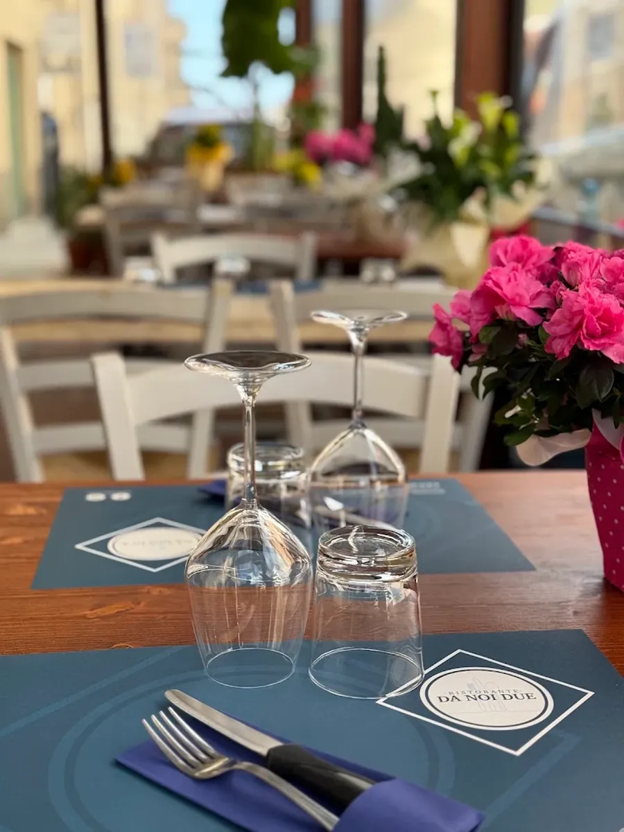 Tavolo apparecchiato con la tovaglietta del ristorante Da Noi Due, bicchieri e fiori, con i tavoli esterni sullo sfondo